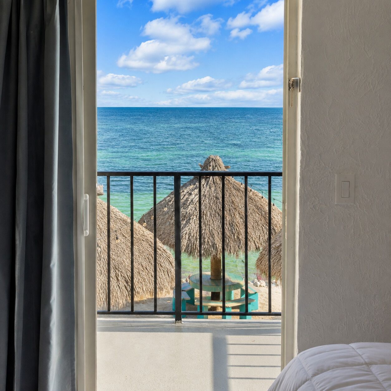 A room with a balcony overlooking a beach with thatched umbrellas and a clear blue sky.