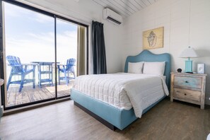 A bedroom with a bed, a nightstand, a lamp, a wall-mounted air conditioner, and a view of a deck with chairs.