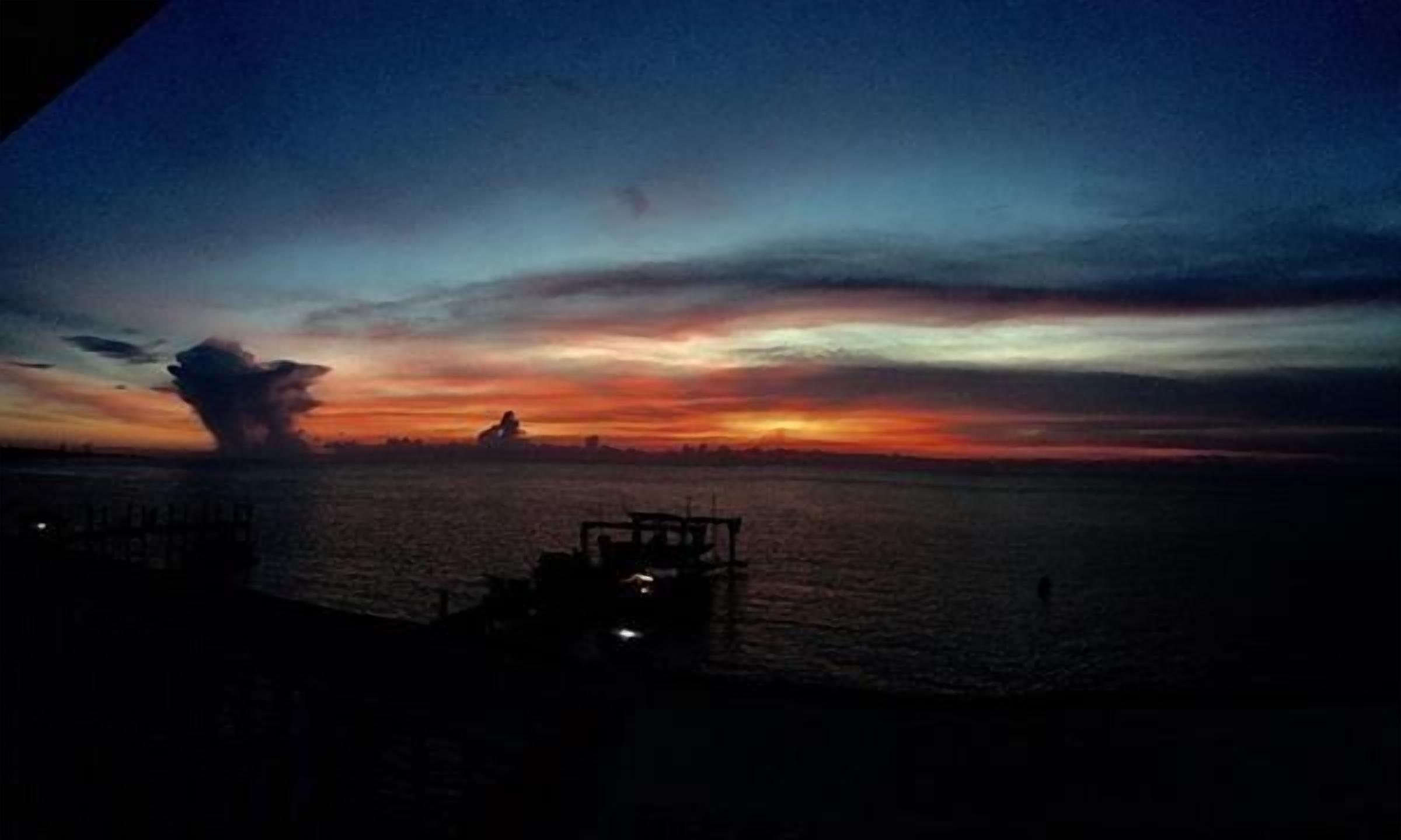 Pôr do sol sobre a água, com a costa distante e uma pequena construção à esquerda.