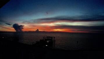 Pôr do sol sobre a água, com a costa distante e uma pequena construção à esquerda.