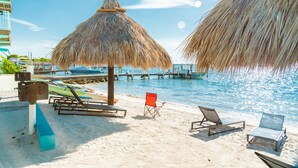 A beach with a thatched umbrella, lounge chairs, and a pool.