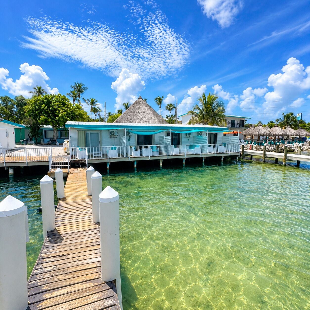 A clear water view leading to a bungalow-style accommodation with a thatched roof, surrounded by palm trees and clear skies.