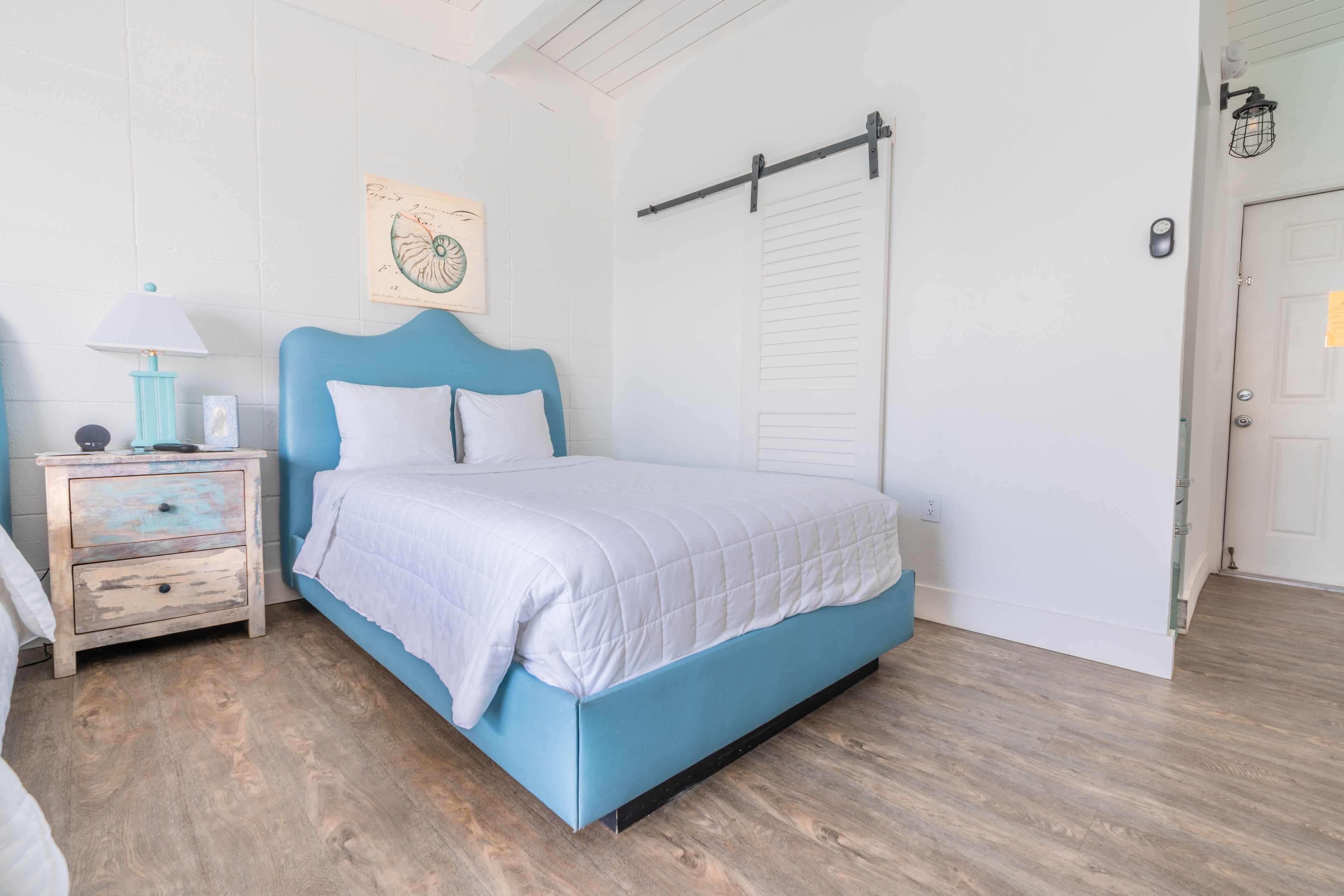 Een slaapkamer met een blauw bed, witte beddengoed, een houten nachtkastje met een lamp en een aan de muur hangend kunstwerk.