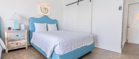 Een slaapkamer met een blauw bed, witte beddengoed, een houten nachtkastje met een lamp en een aan de muur hangend kunstwerk.