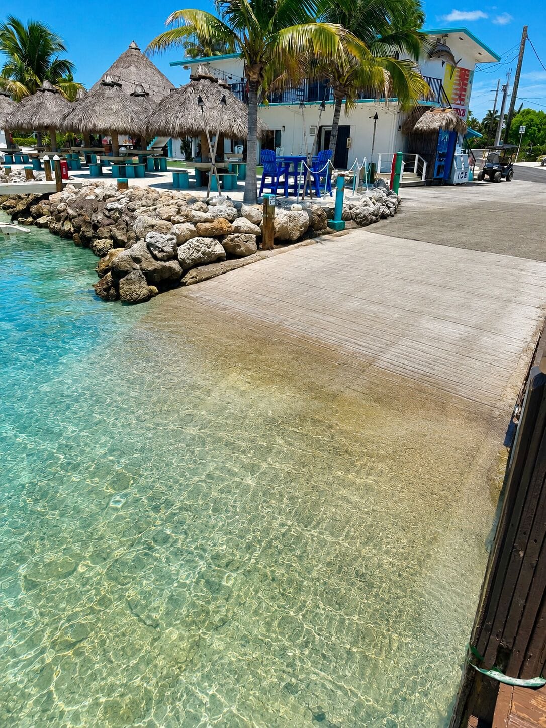 A clear tropical pool with a rocky edge, a thatched-roof building, and a paved walkway.
