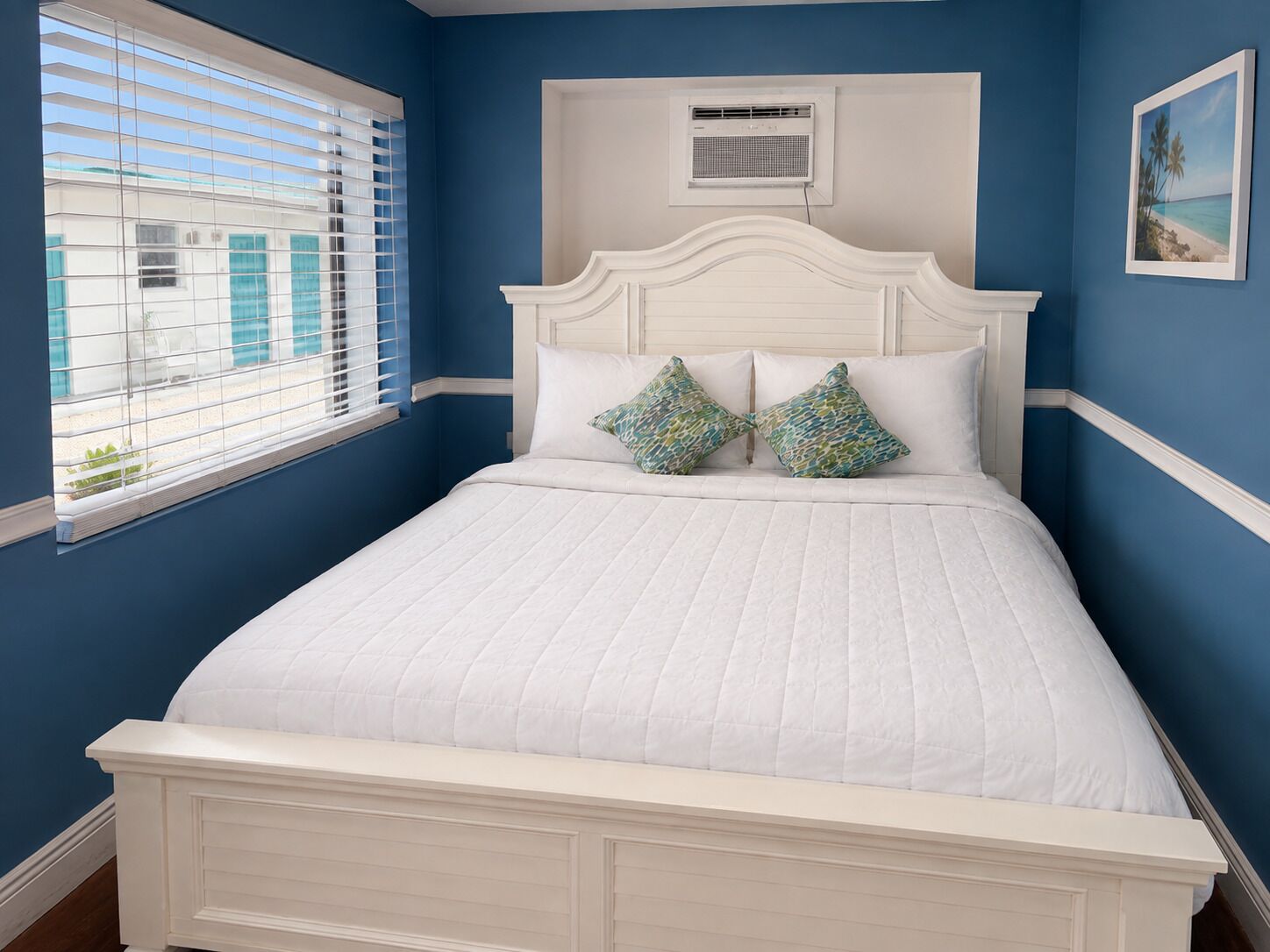 A bedroom with a white bed, two patterned pillows, a window with blinds, and a wall-mounted air conditioning unit.