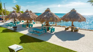 A beachside area with thatched umbrellas, picnic tables, and a pool.