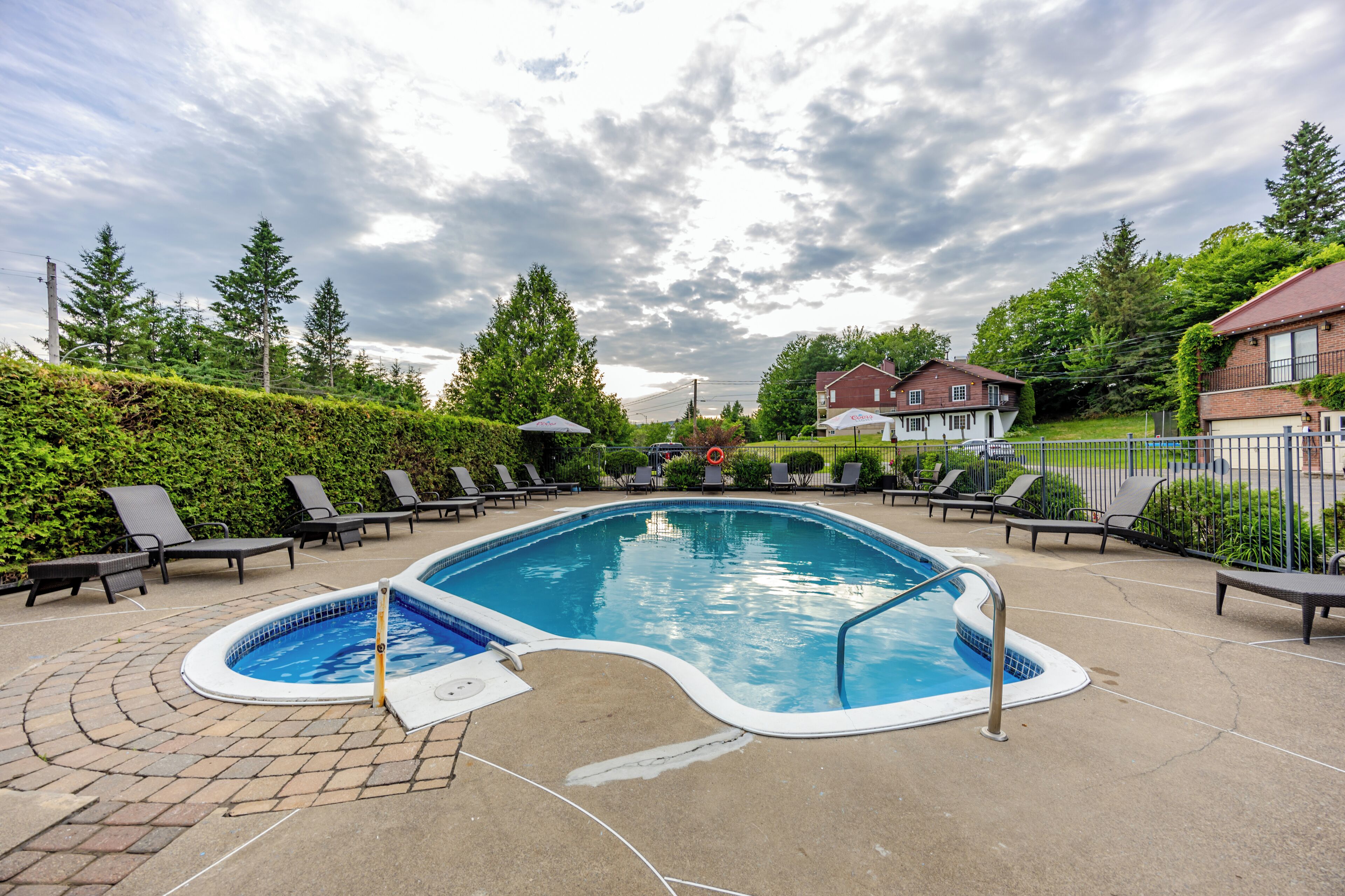 A swimming pool with lounge chairs and a hot tub, surrounded by greenery and houses.