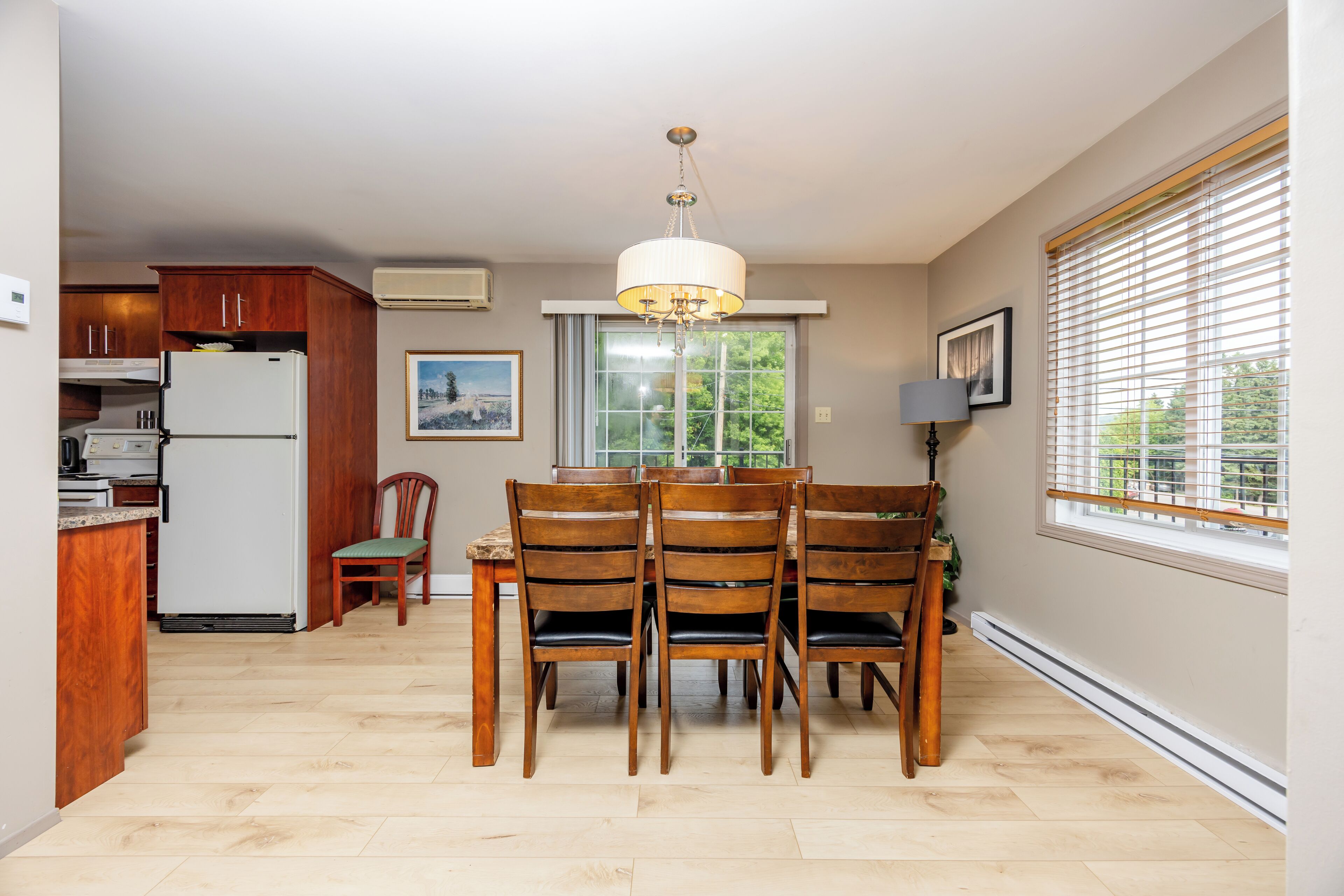 A kitchen with wooden cabinets, a white refrigerator, a dining table with chairs, a chandelier, and a window with blinds.