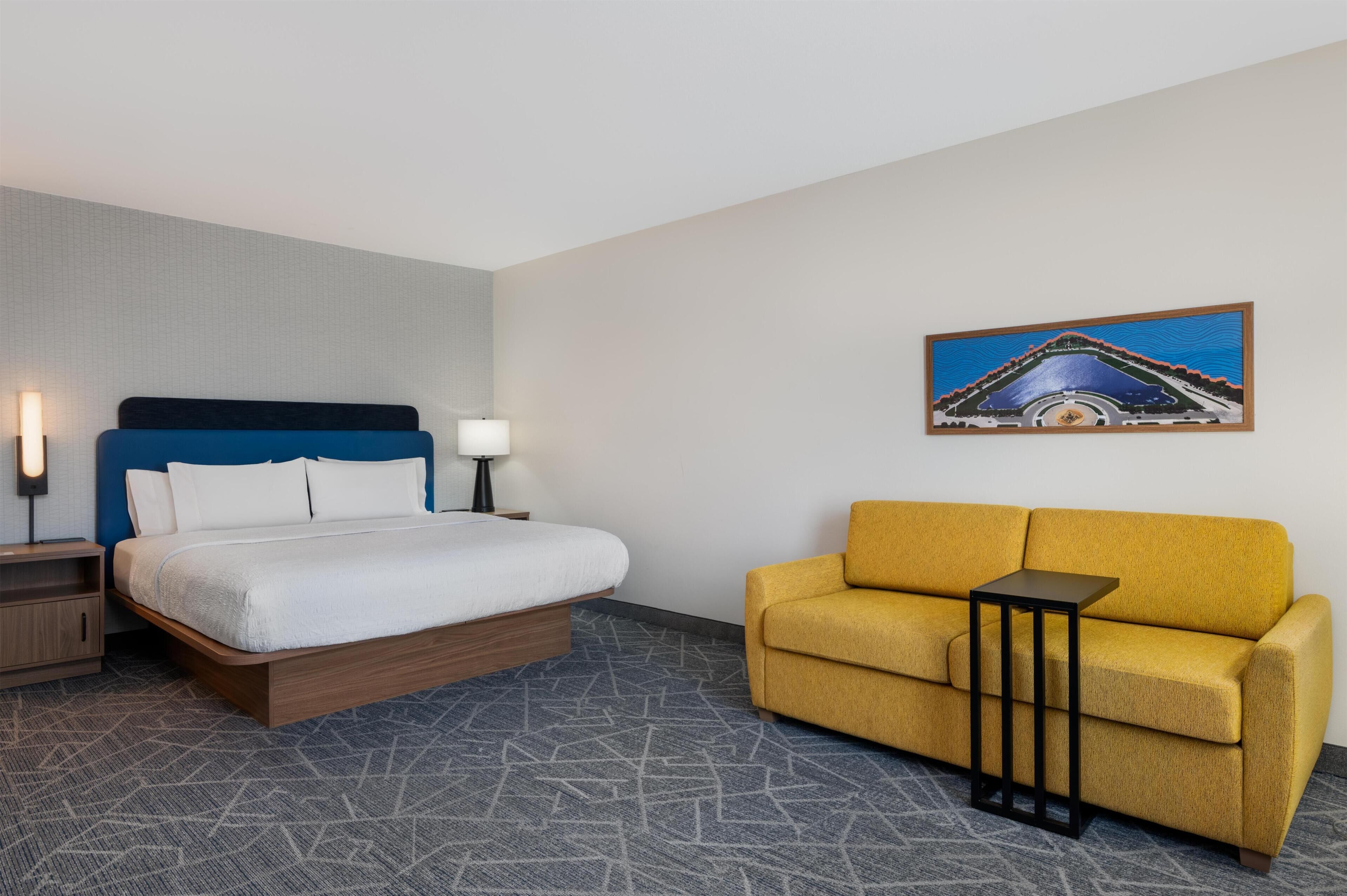 A hotel room with a bed, a sofa, a small table, and a painting on the wall.