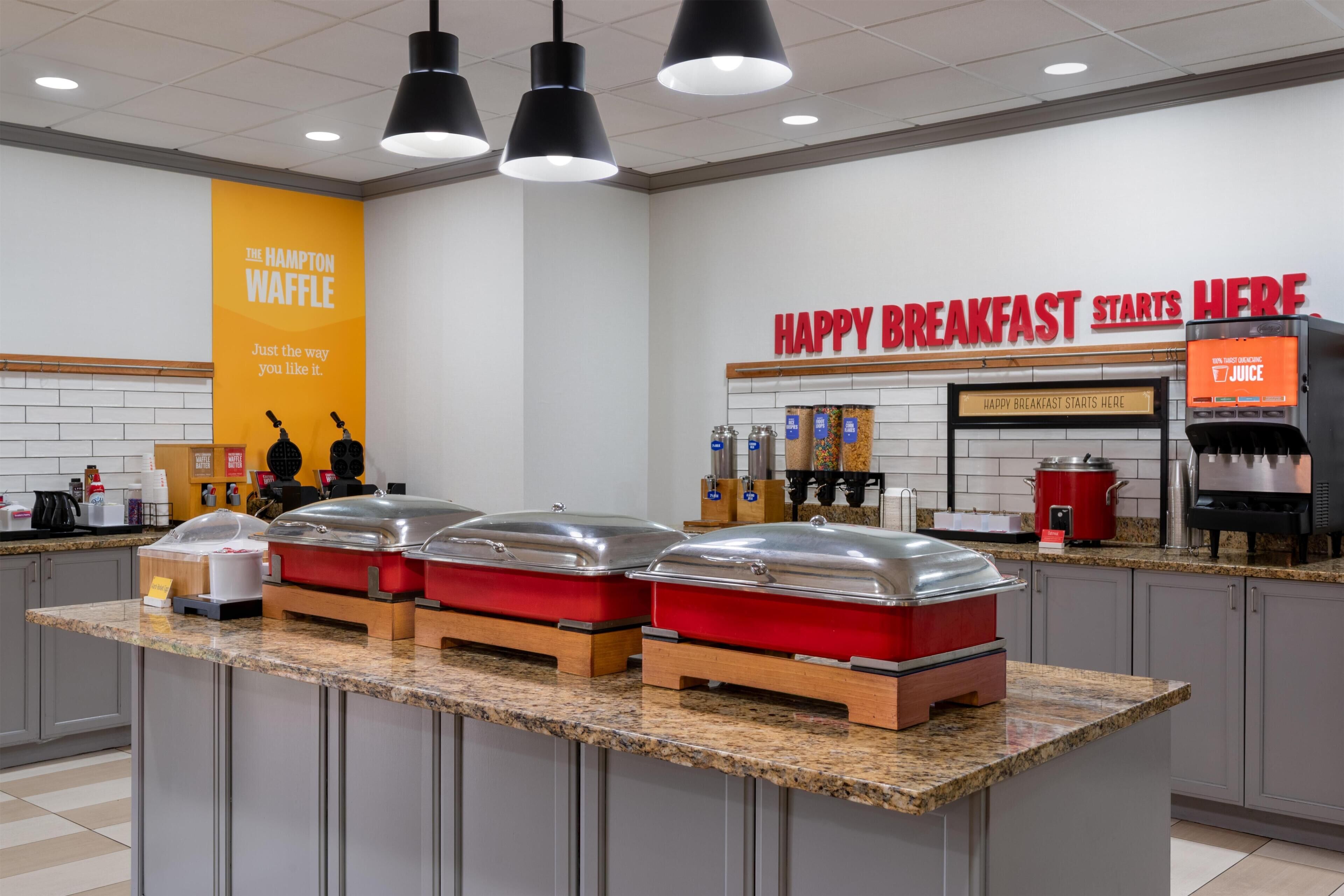 A breakfast buffet area with a variety of food options, a juice dispenser, and a sign indicating the location for breakfast.