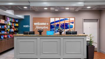 A hotel reception area with a 'Welcome to Hampton' sign, a 'Treats' section with snacks, and a 'Hilton Honors' loyalty program sign.