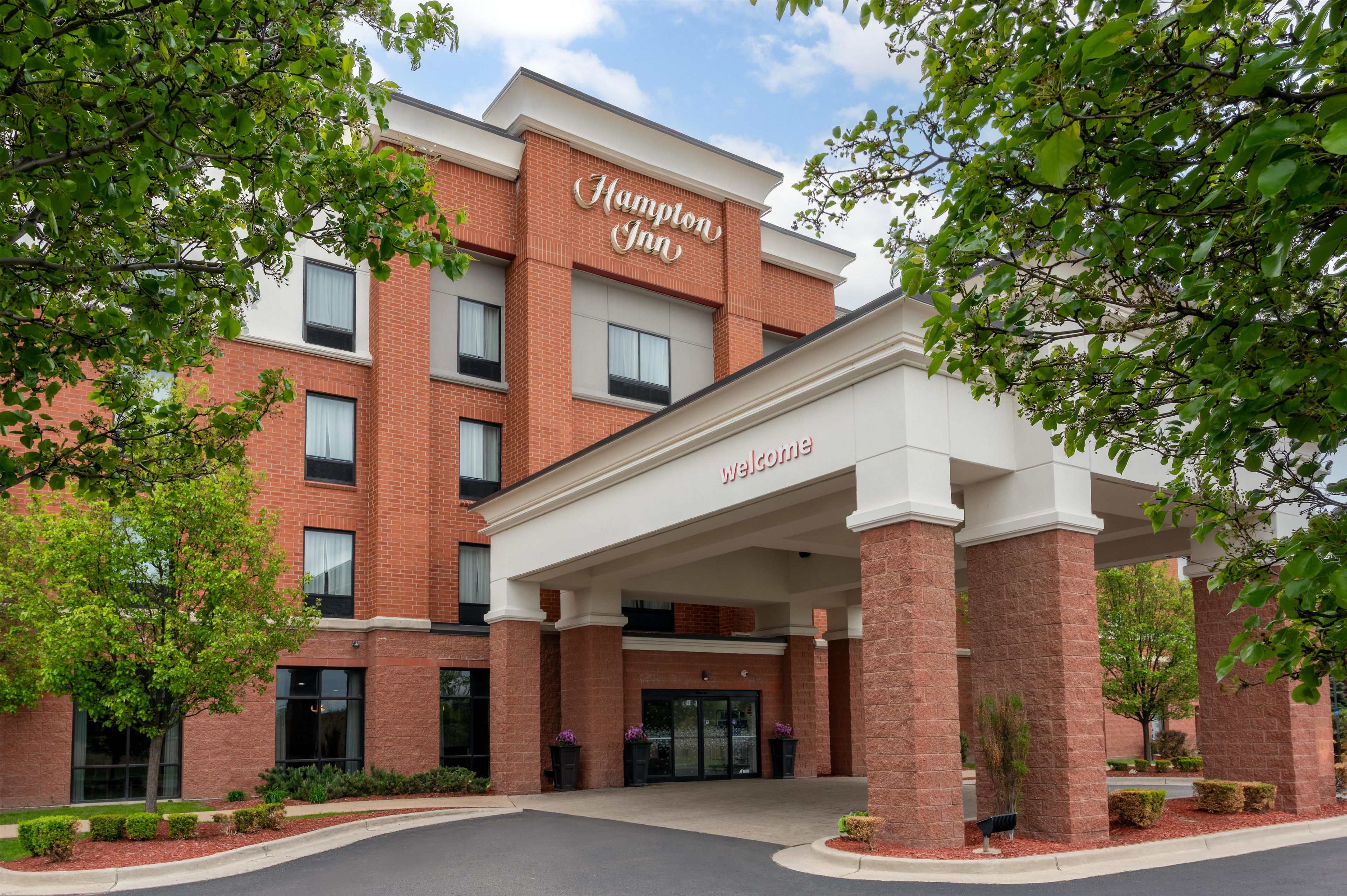 A brick building with a 'Welcome' sign, likely a hotel, with a covered entrance and surrounding greenery.