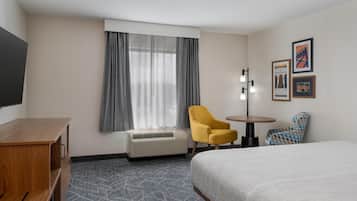 A hotel room with a bed, a desk, a chair, a table, an air conditioner, and a window with curtains.
