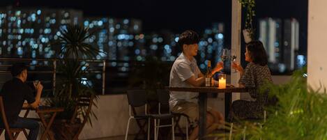 A rooftop terrace with tables and chairs, a city skyline in the background, and a decorative lamp.