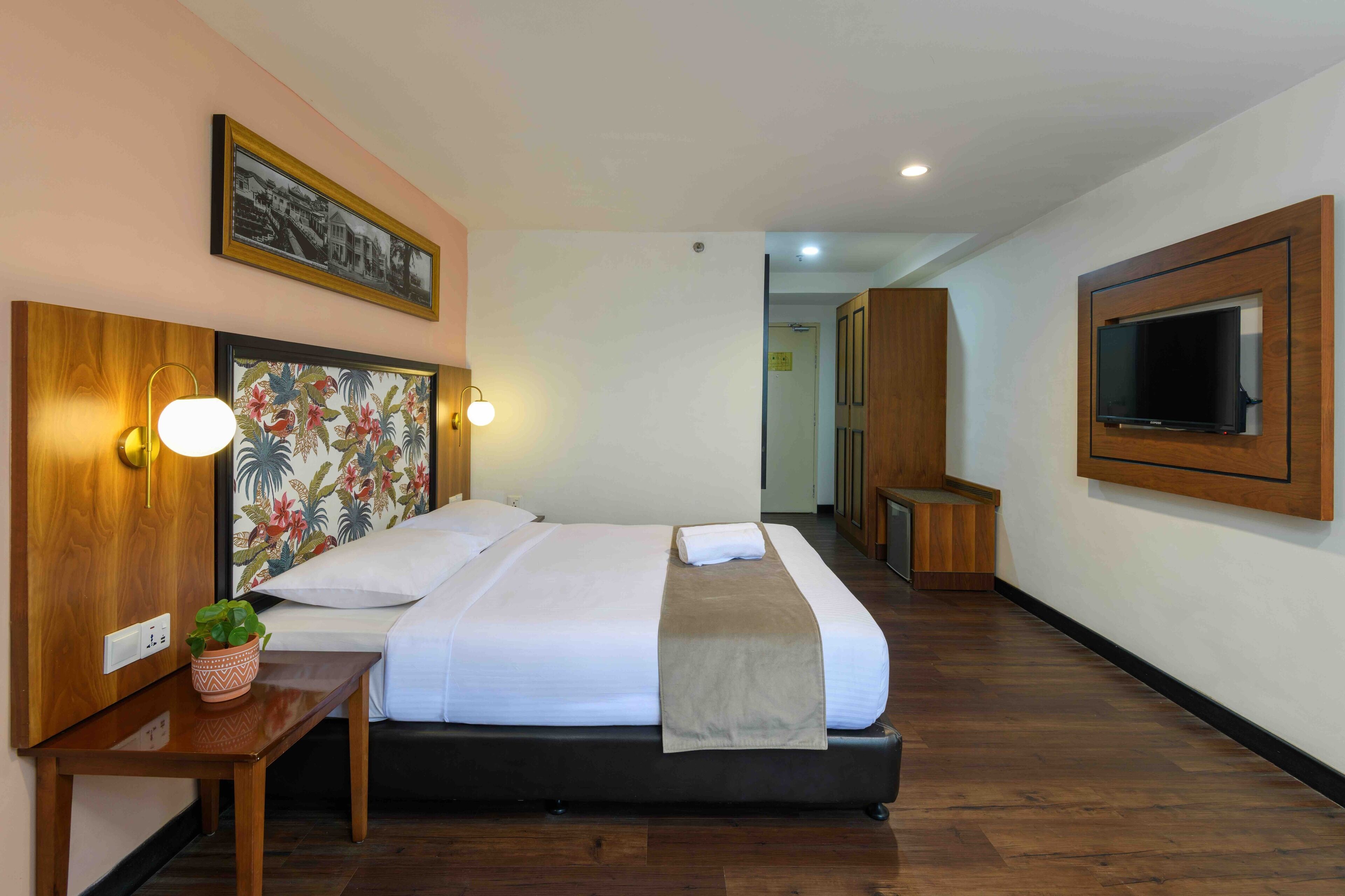 A hotel room with a large bed, a wooden headboard, a wall-mounted TV, and a small table with a plant.