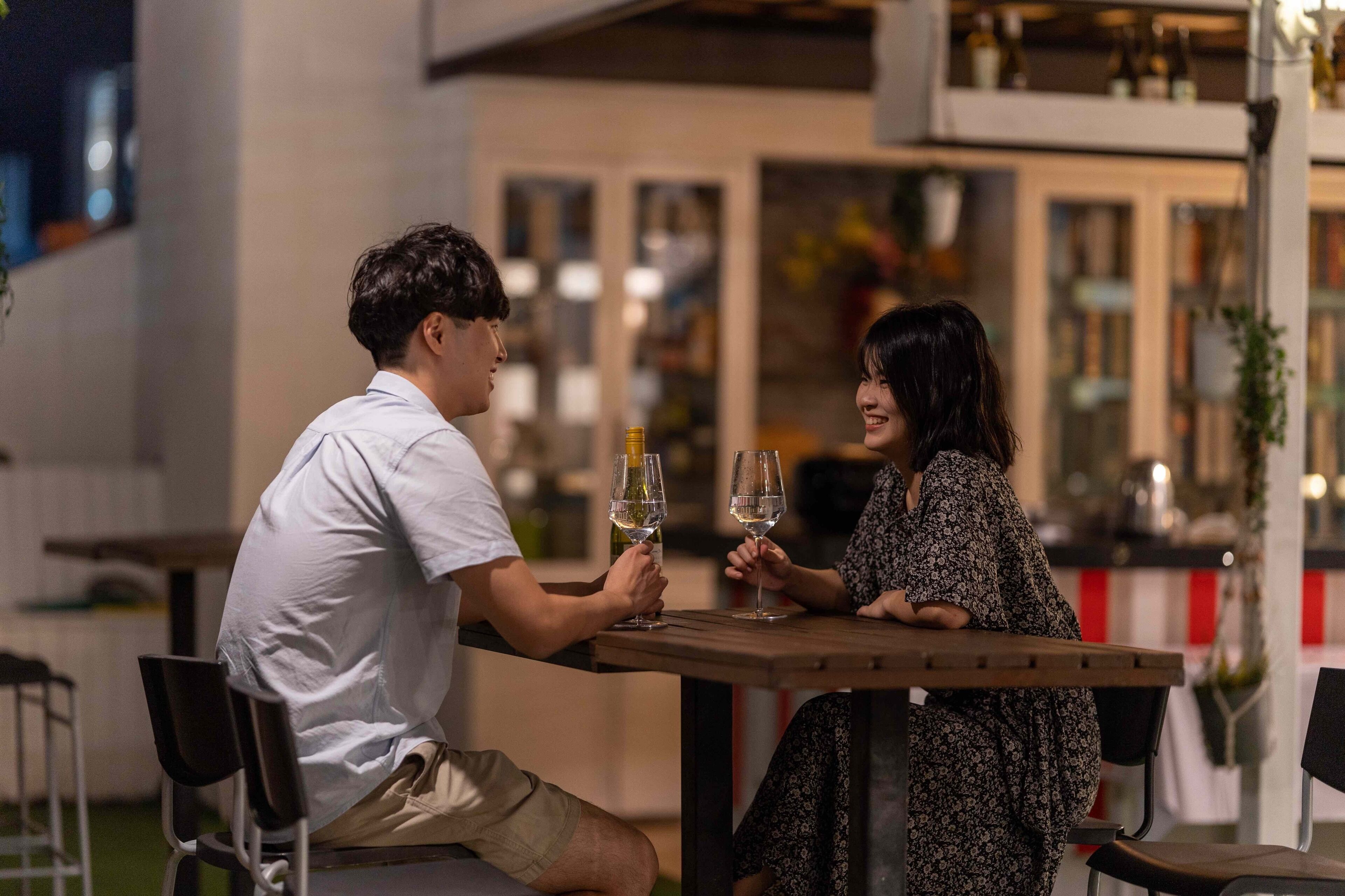 A couple seated at an outdoor table with wine glasses, in a modern setting with a view of a building and indoor seating area.