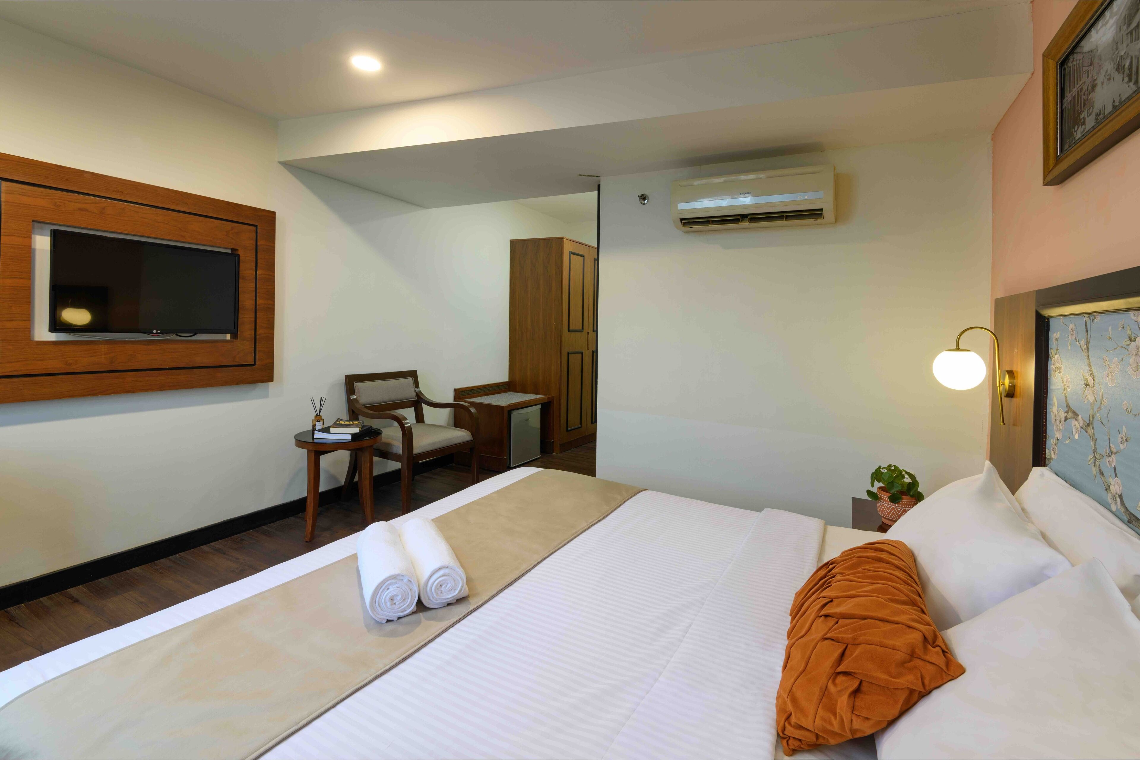 A hotel room with a bed, a TV mounted on the wall, a desk with a chair, and an air conditioning unit.