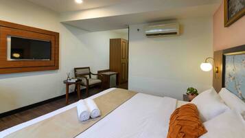 A hotel room with a bed, a TV mounted on the wall, a desk with a chair, and an air conditioning unit.