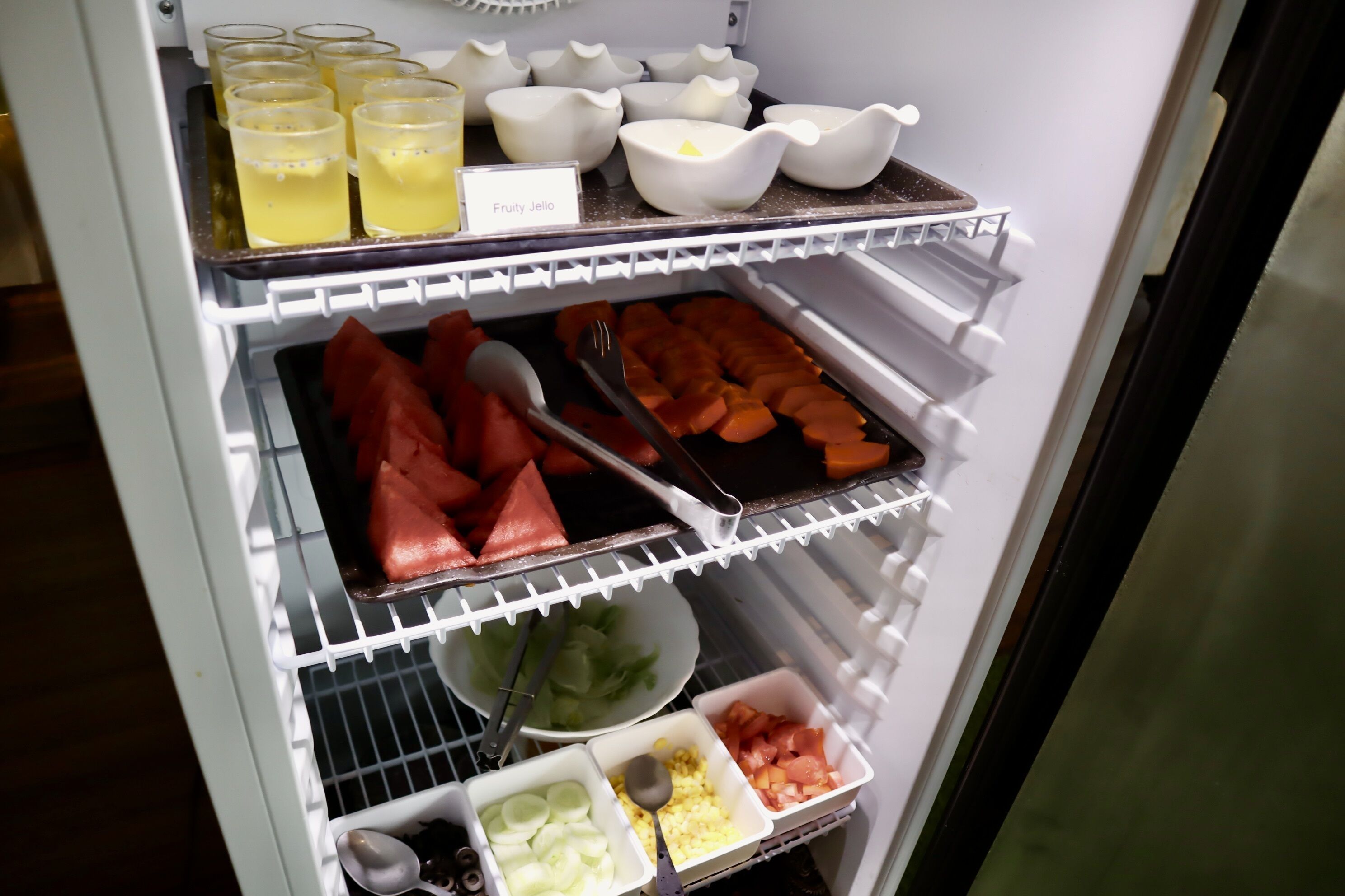 A refrigerator stocked with various food items including watermelon, corn, and jellies.