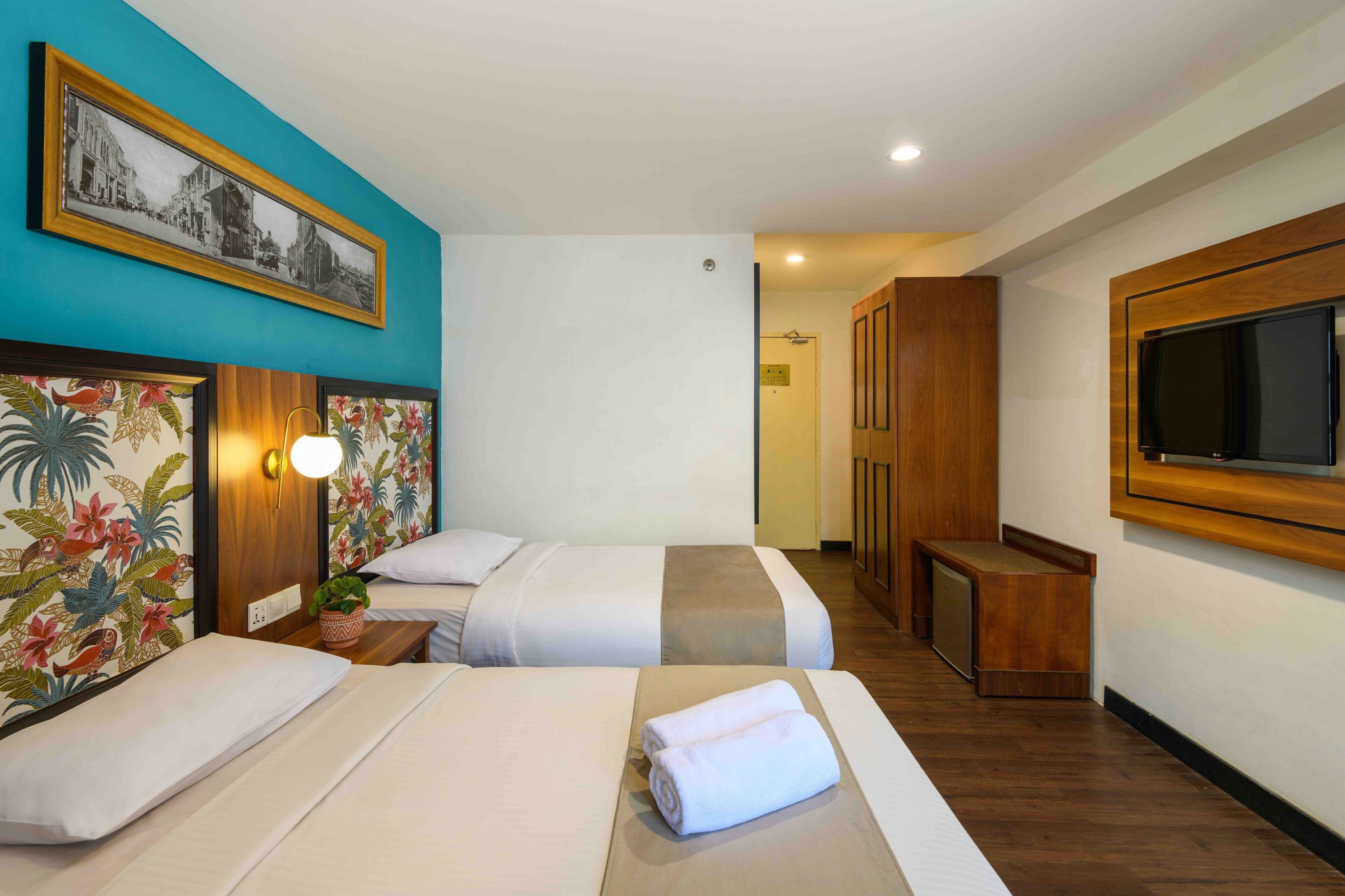 A hotel room with two beds, a TV, a wooden bench, and a wall-mounted TV.