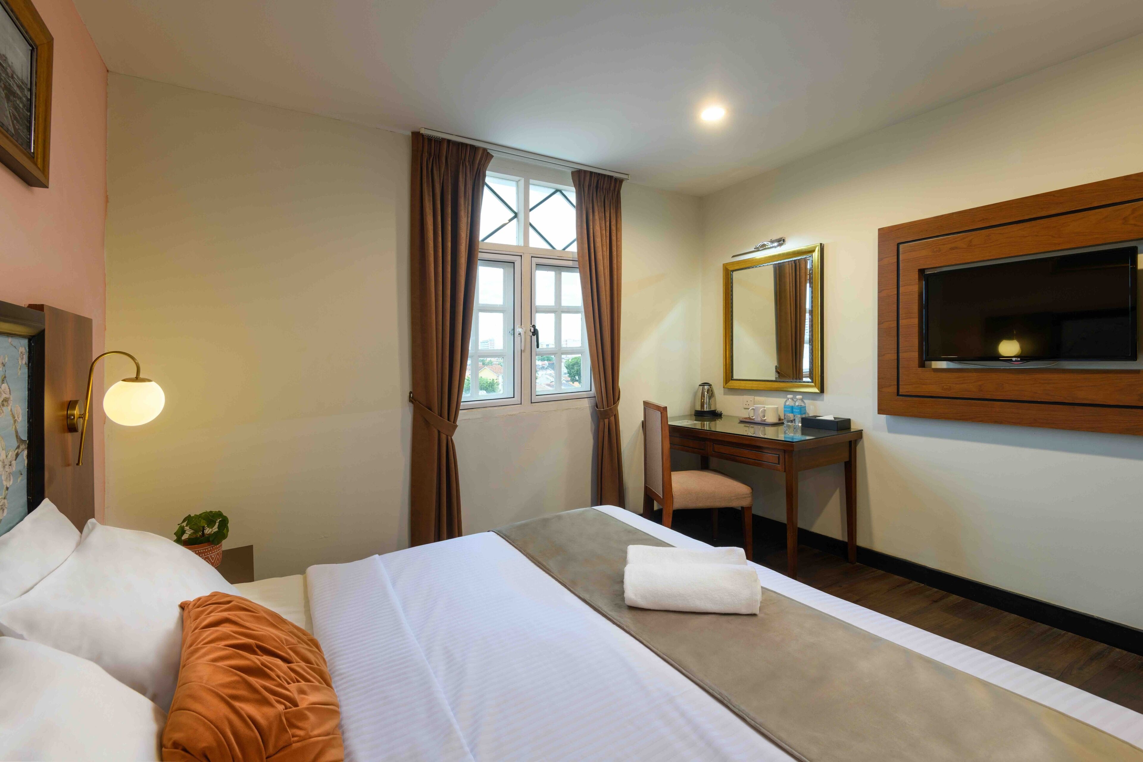 A hotel room with a bed, a desk, a chair, a desk lamp, a mirror, and a window with curtains.