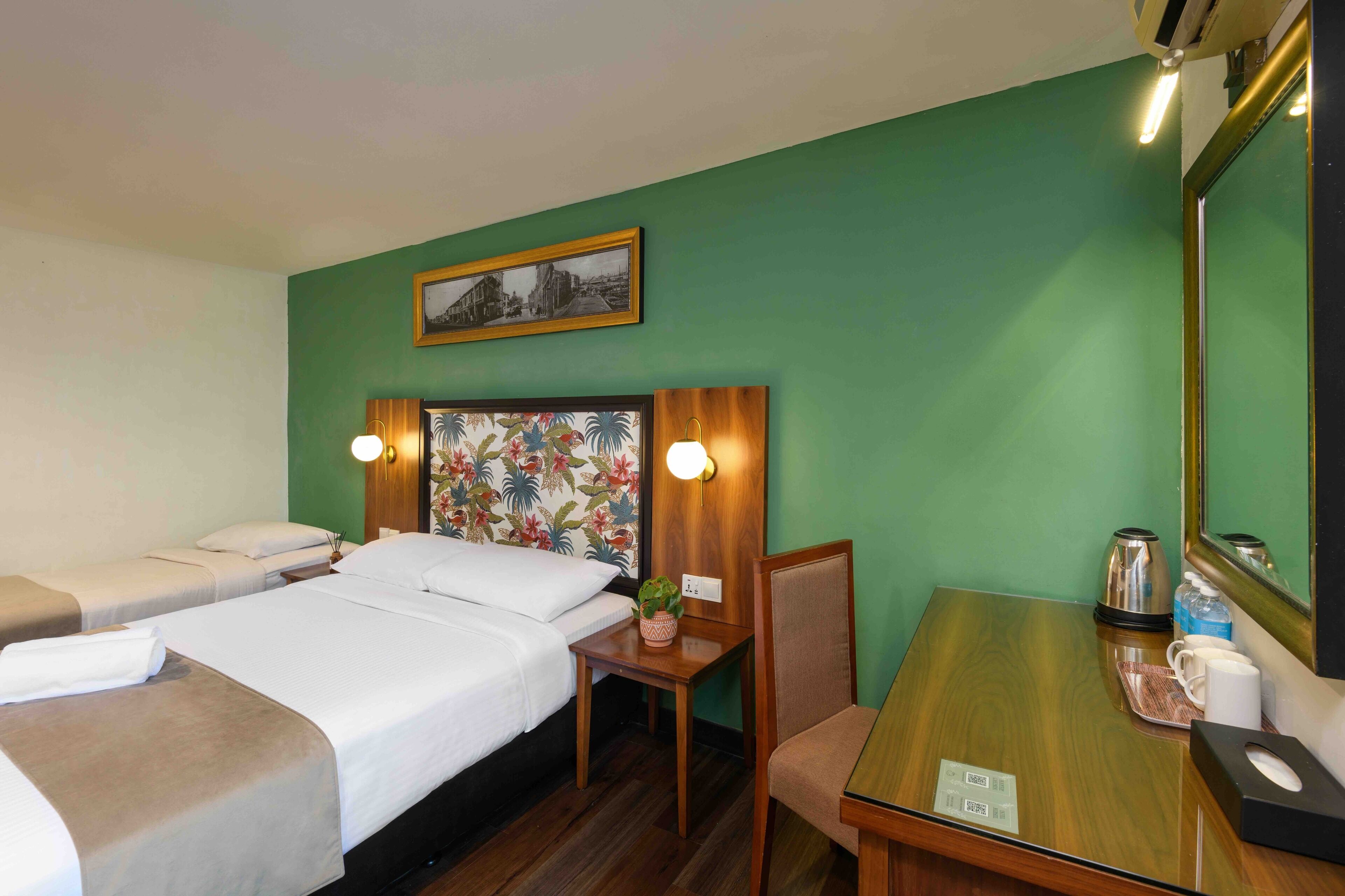 A hotel room with two beds, a wooden desk, a chair, a small table, a mirror, and a wall-mounted picture.