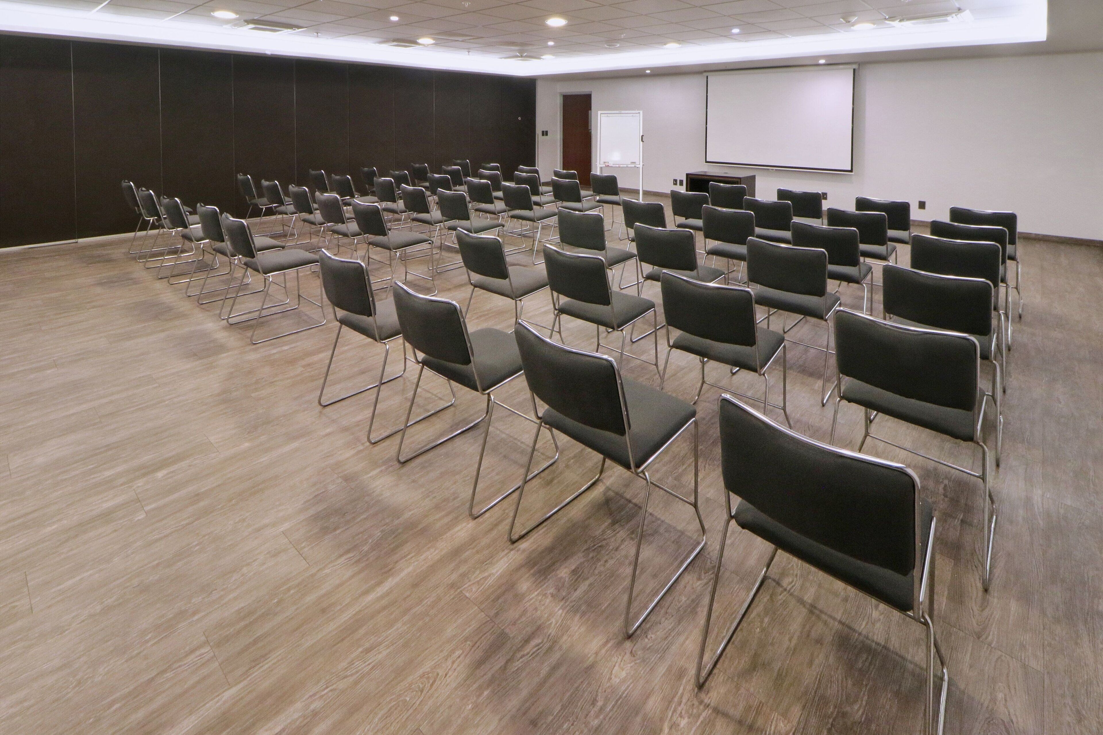 Un salón de conferencias con filas de sillas, una pizarra y una pantalla de proyección.