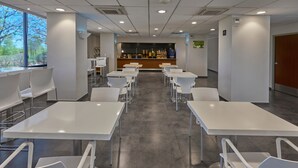 Una sala de conferències moderna amb taules i cadires de color blanc, una zona de recepció amb mostrador i vistes a arbres a l'exterior.