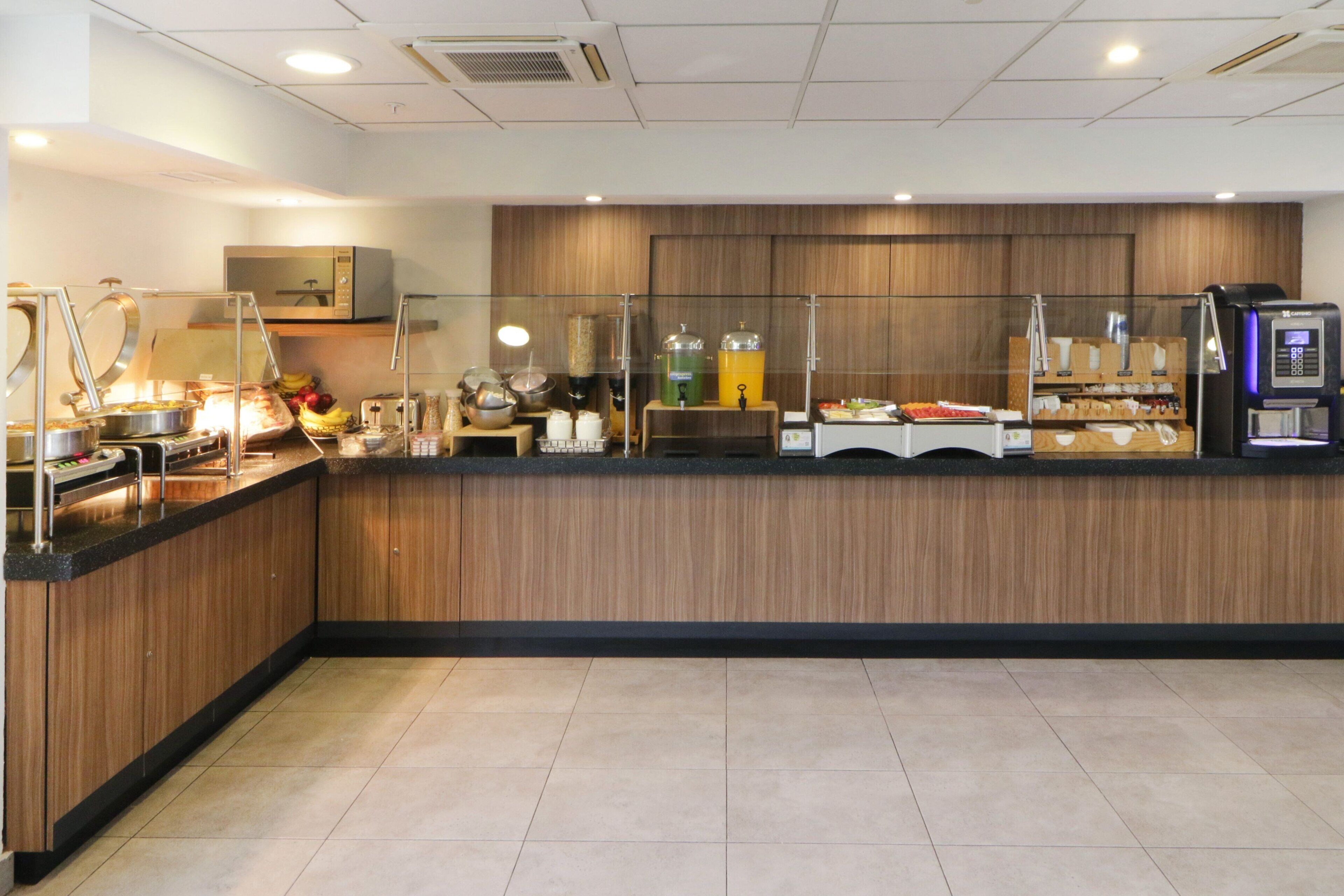 Una barra de desayuno moderna con detalles de madera, equipada con una máquina de café, dispensadores de jugo y diferentes opciones de alimentos.