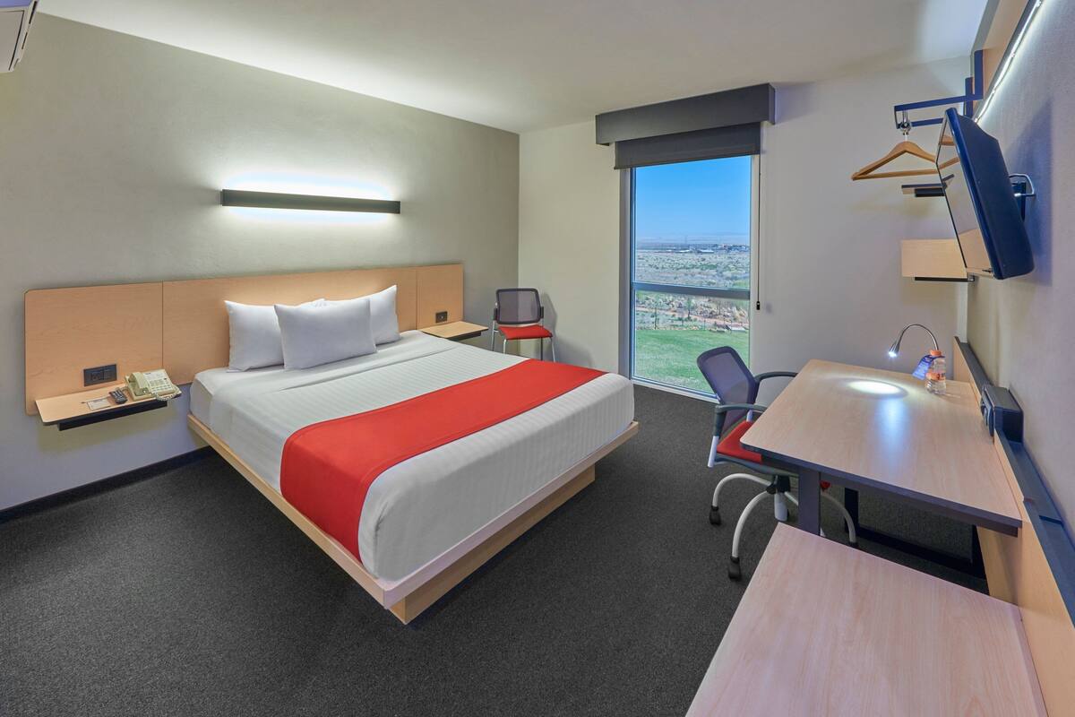 A hotel room with a bed, bedside table, chair, desk, and a large window offering a city view.