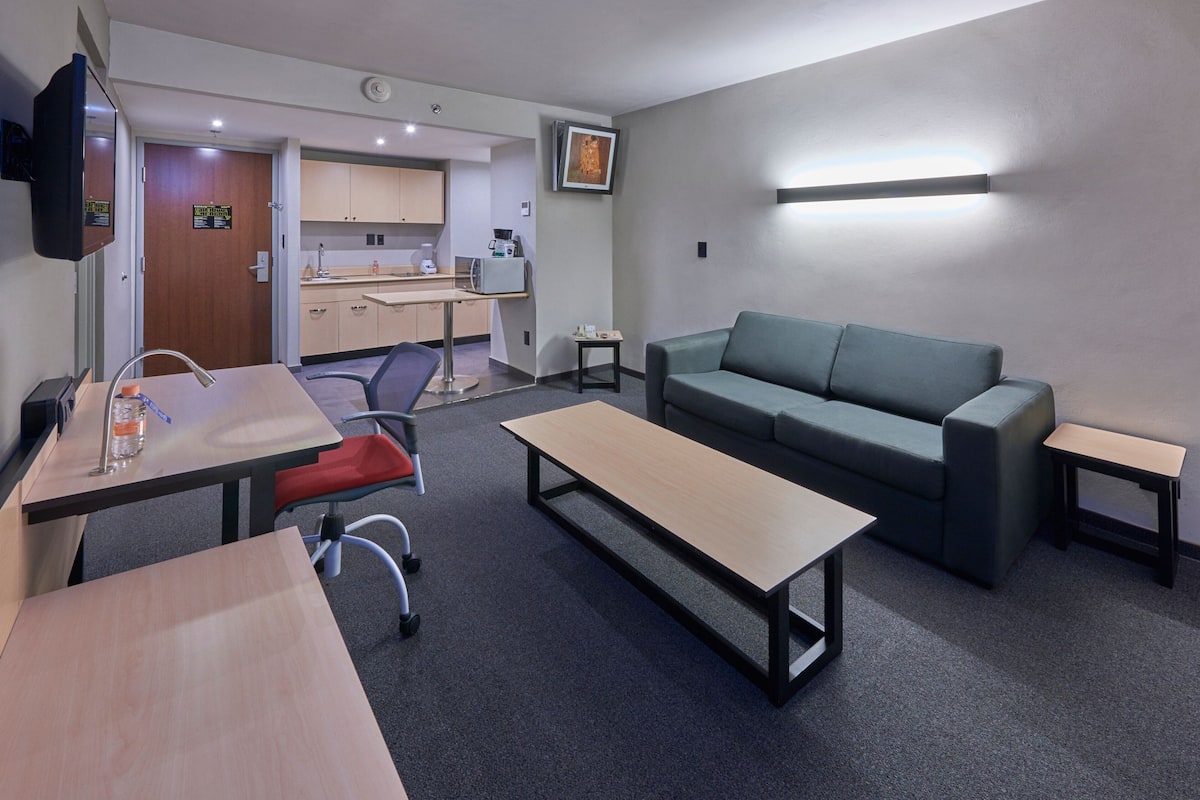 A modern hotel room with a kitchenette, a sofa, a dining table, and a TV.