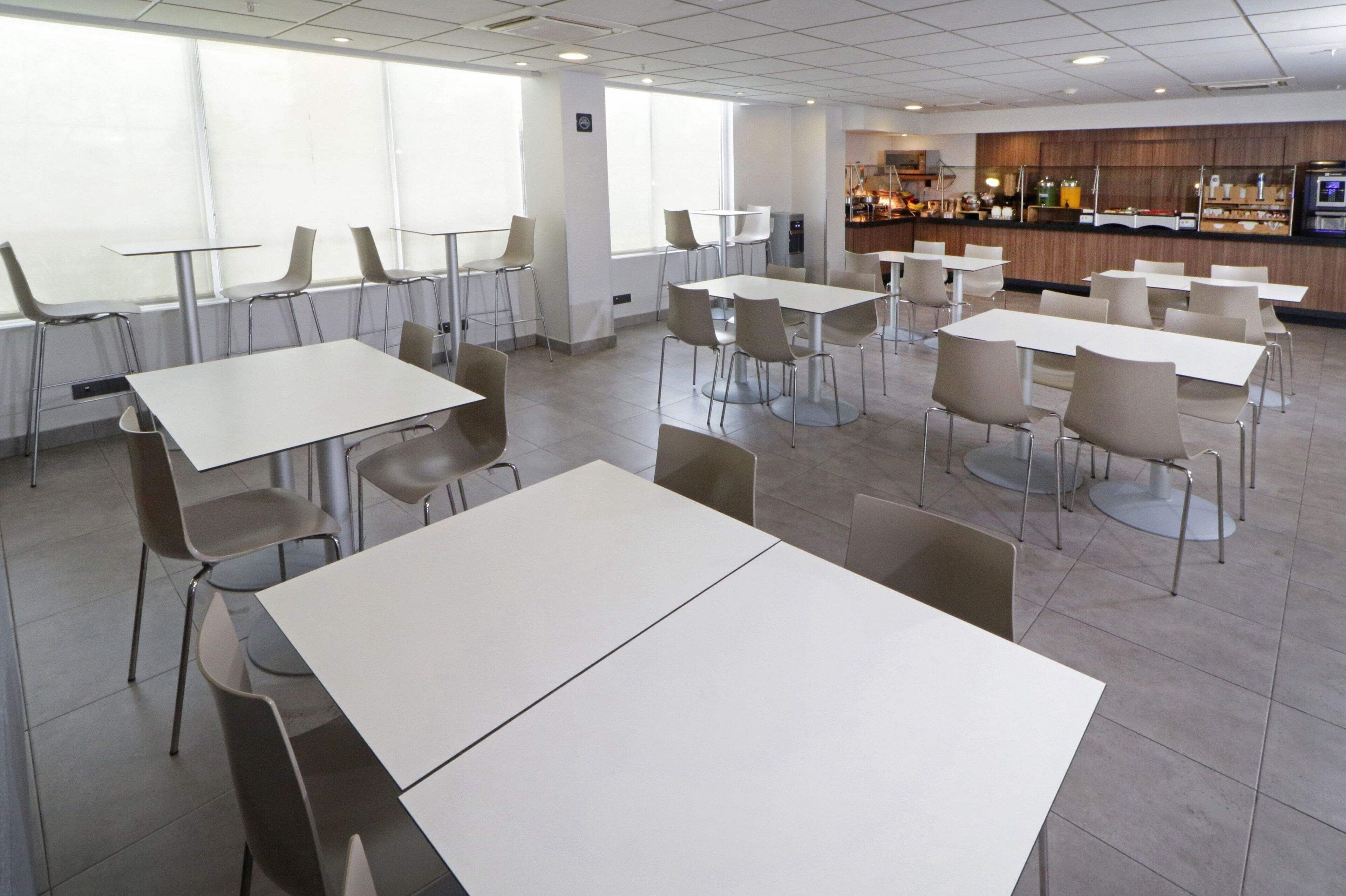 A spacious cafeteria with multiple tables and chairs, large windows, and a counter area with food and beverages.