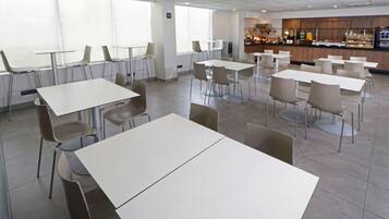A spacious cafeteria with multiple tables and chairs, large windows, and a counter area with food and beverages.