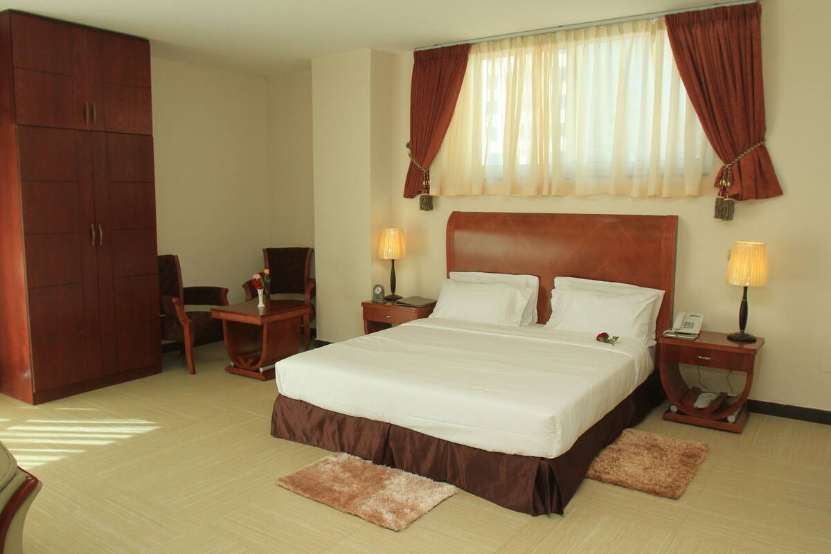 A hotel room with a large bed, two bedside tables with lamps, a wardrobe, and a desk with a chair.