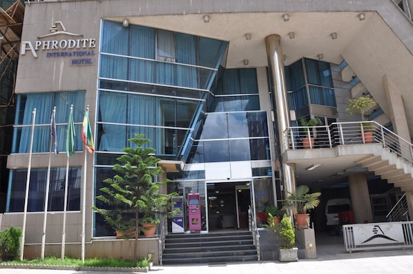 The exterior of the Aphrodite International Hotel with a modern design, featuring a staircase leading to the entrance, multiple flags, and a visible ATM machine.