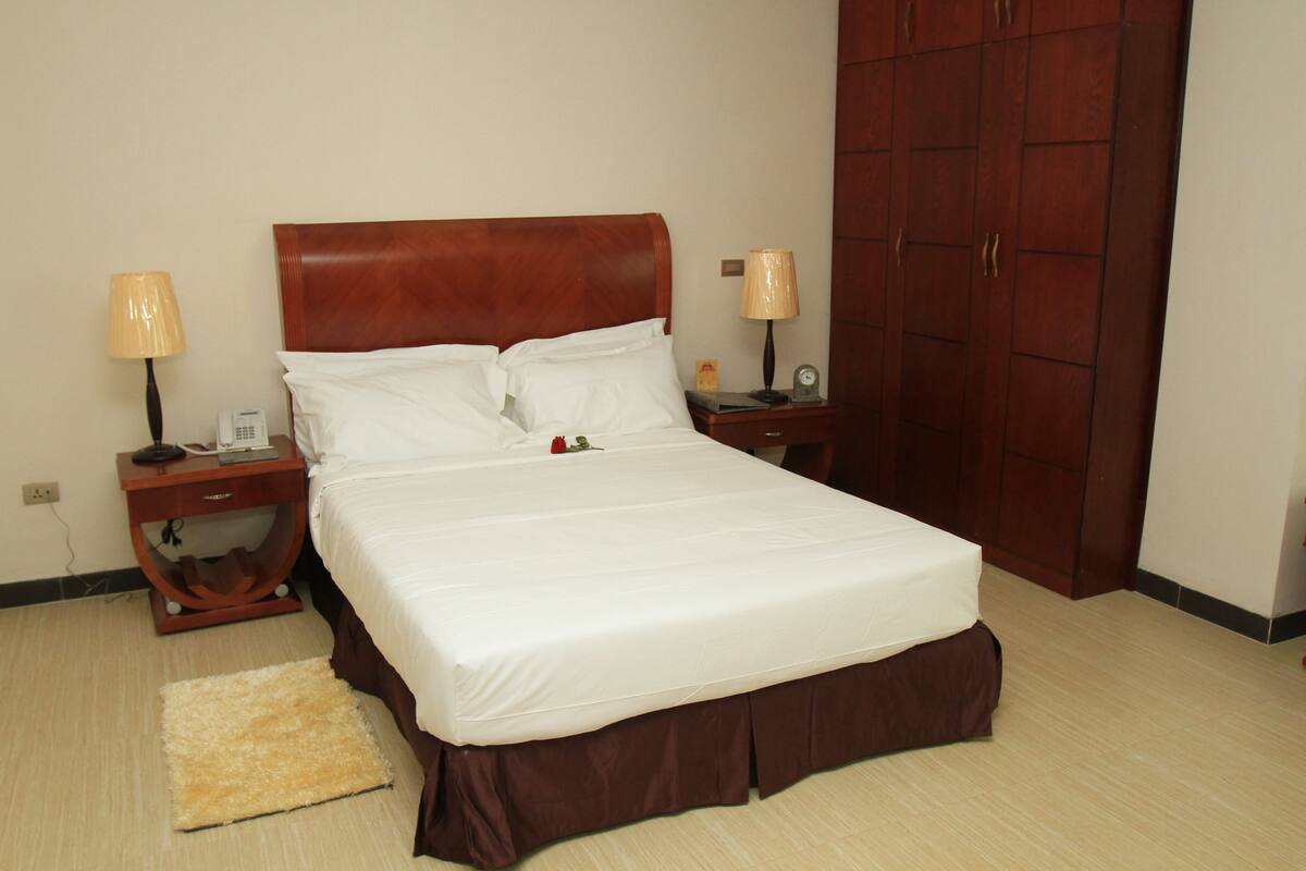 A neatly made bed with white linens, two bedside lamps, a nightstand, a wooden wardrobe, and a small rug on a light-colored floor.