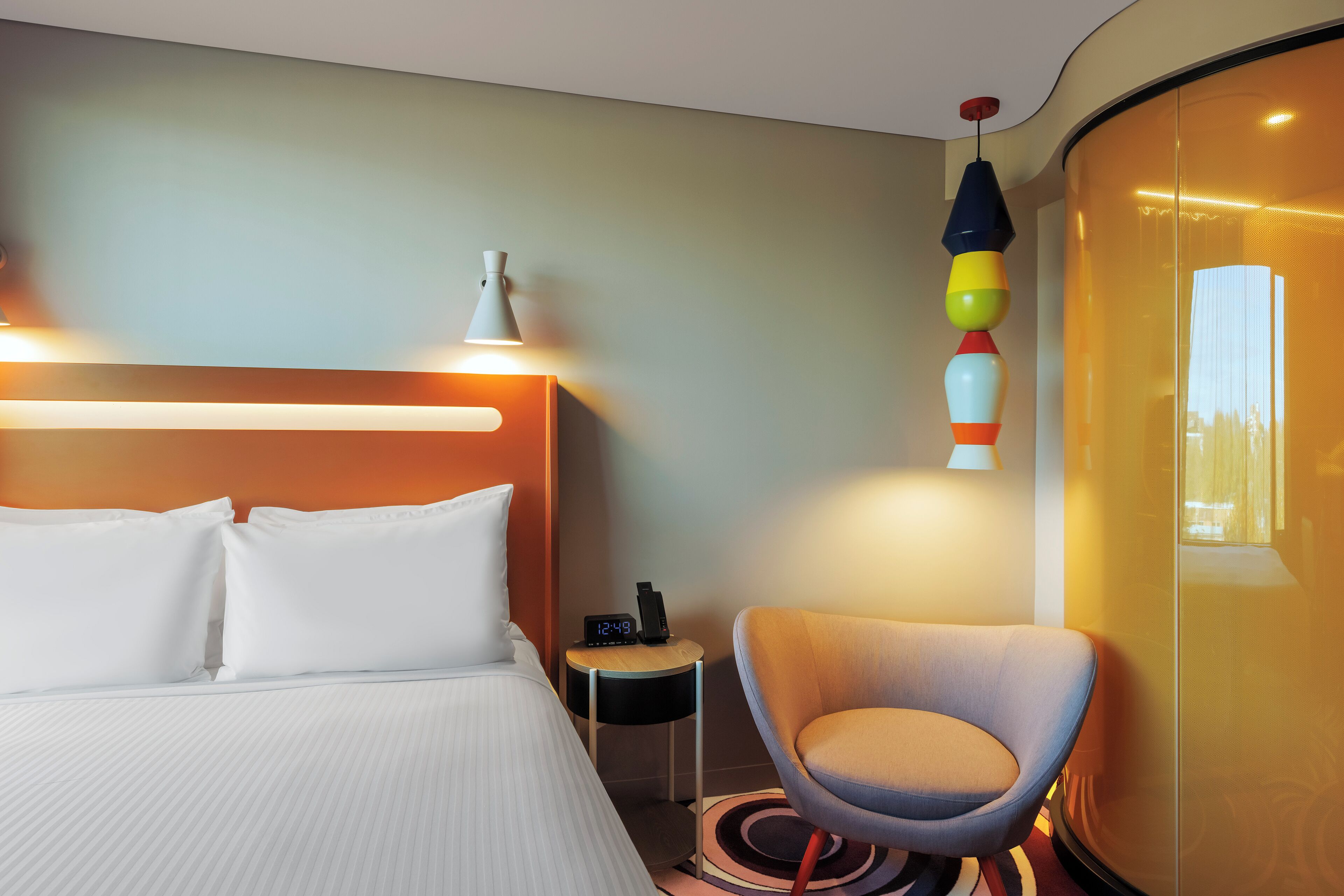 A hotel room with a bed, a chair, a bedside table with a clock, and a wall-mounted lamp.