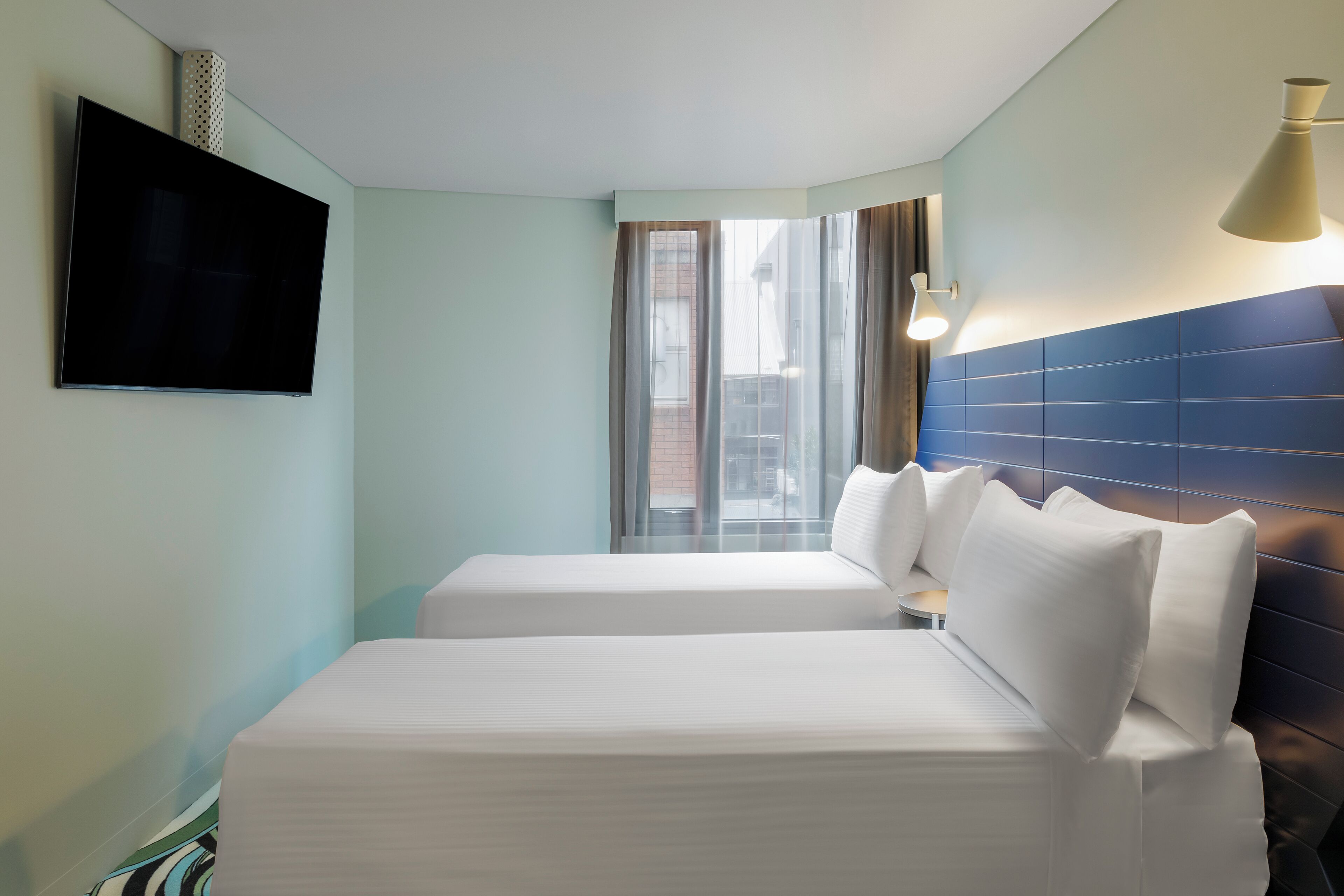 A hotel room with two beds, a flat-screen TV, a window with curtains, and a blue-tiled headboard.