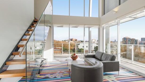 A modern living room with a staircase, large windows, and a colorful striped rug. - ADGE Hotel & Residences (Surry Hills)