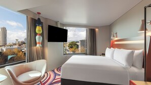 A hotel room with a large bed, a chair, a TV, and a view of the city. - ADGE Hotel & Residences (Surry Hills)