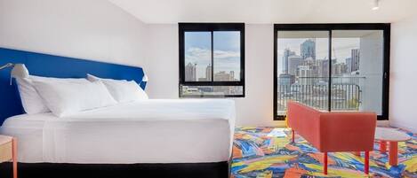 A hotel room with a large bed, a city view through a sliding door, a colorful abstract floor, and a red chair.
