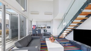 A modern living room with a sofa, a coffee table, and a TV. There is a staircase with glass railings and wooden steps. - ADGE Hotel & Residences (Surry Hills)