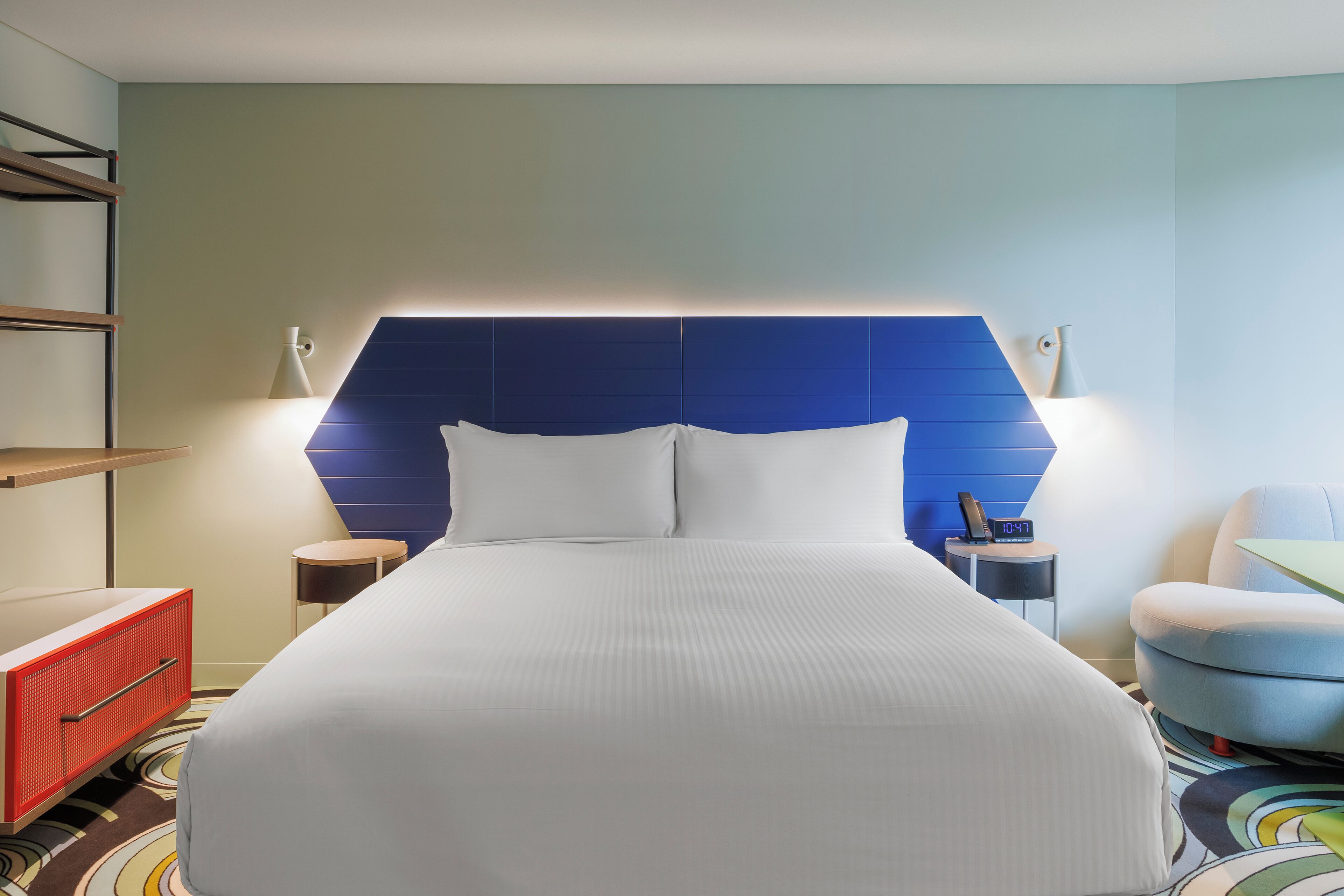 Une chambre d’hôtel avec un grand lit, un meuble de rangement rouge, une tête de lit bleue, deux tables de chevet avec des lampes et une petite table ronde.