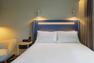 A hotel room with a bed, bedside table, lamp, chair, and a clock.