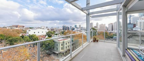 Un balcón en un edificio alto con vista a la ciudad, barandas de vidrio y piso de baldosas.