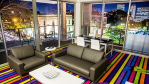A modern living room with a sofa, chairs, and a coffee table. Large windows offer a city view. - ADGE Hotel & Residences (Surry Hills)