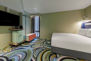 A hotel room with a bed, a TV, a minibar, a shower, and a colorful carpet.