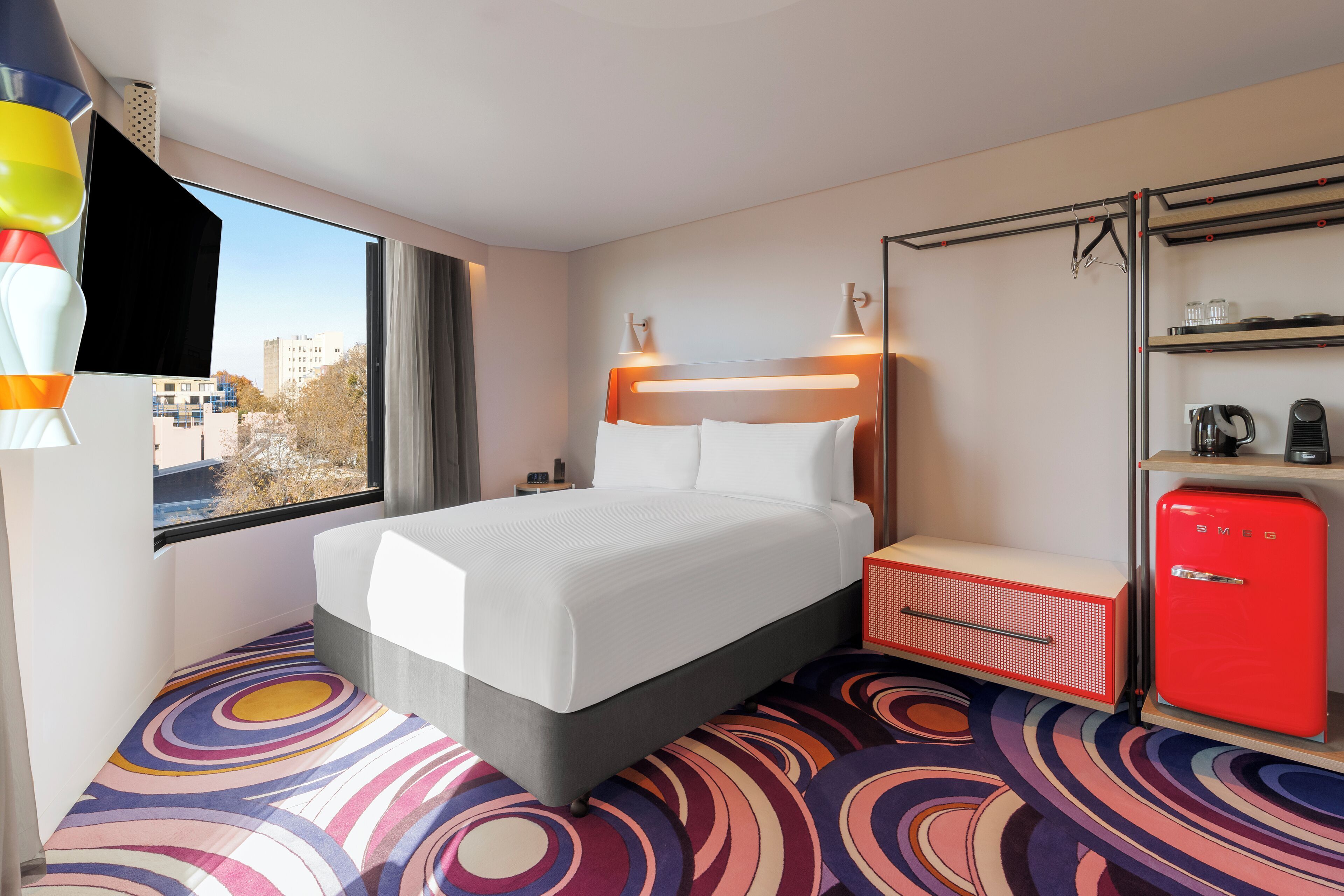 A hotel room with a bed, a small red refrigerator, a shelf with items, a window with curtains, and a view of buildings outside.