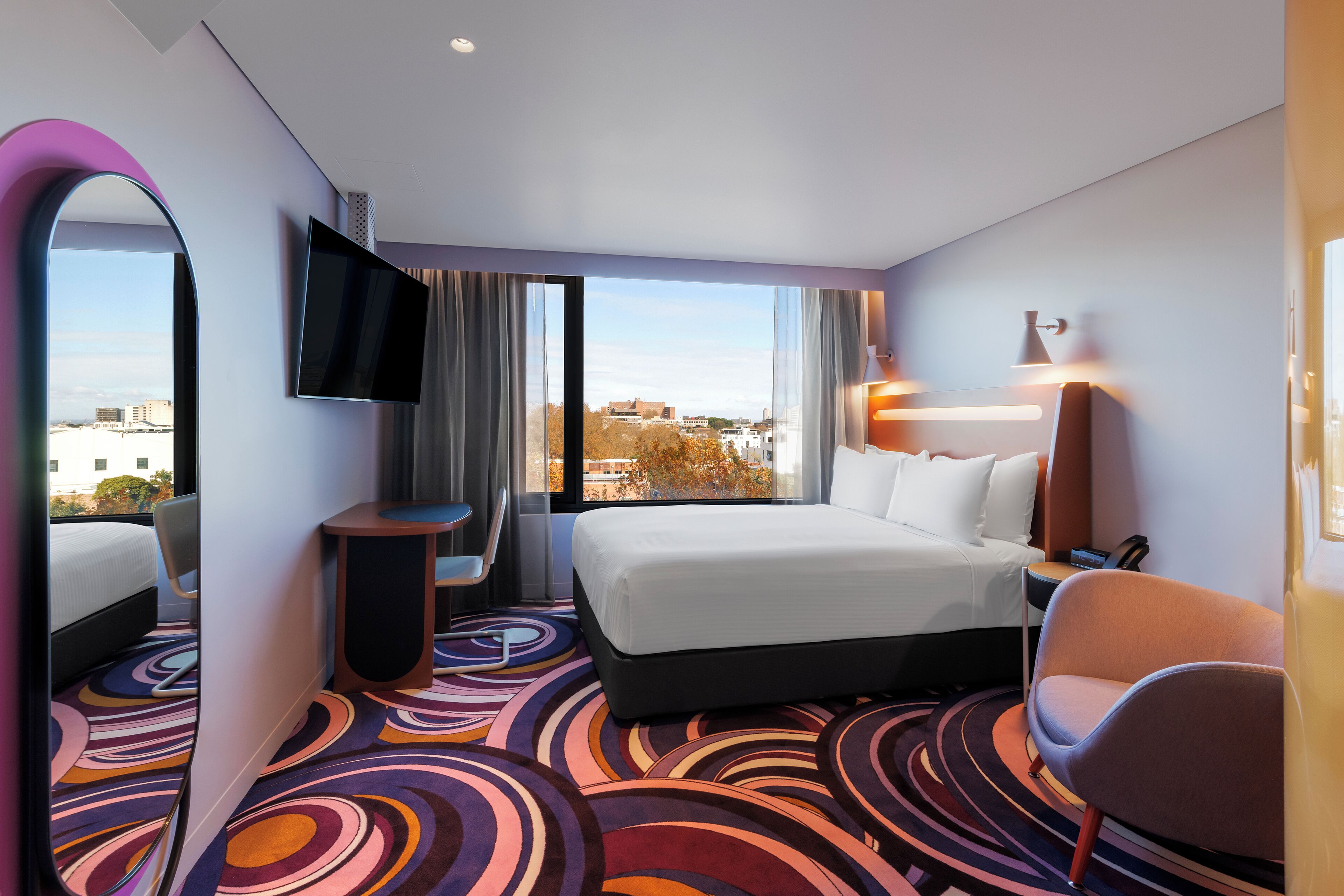 A hotel room with a large bed, a TV, a desk, and a view of the city.